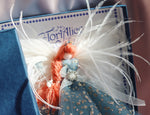 Load image into Gallery viewer, A close-up of a decorative doll or ornament inside a blue gift box. The figure has long white feathers, orange yarn hair, a light blue dress with small gold floral details, and a jeweled ornament at the waist.
