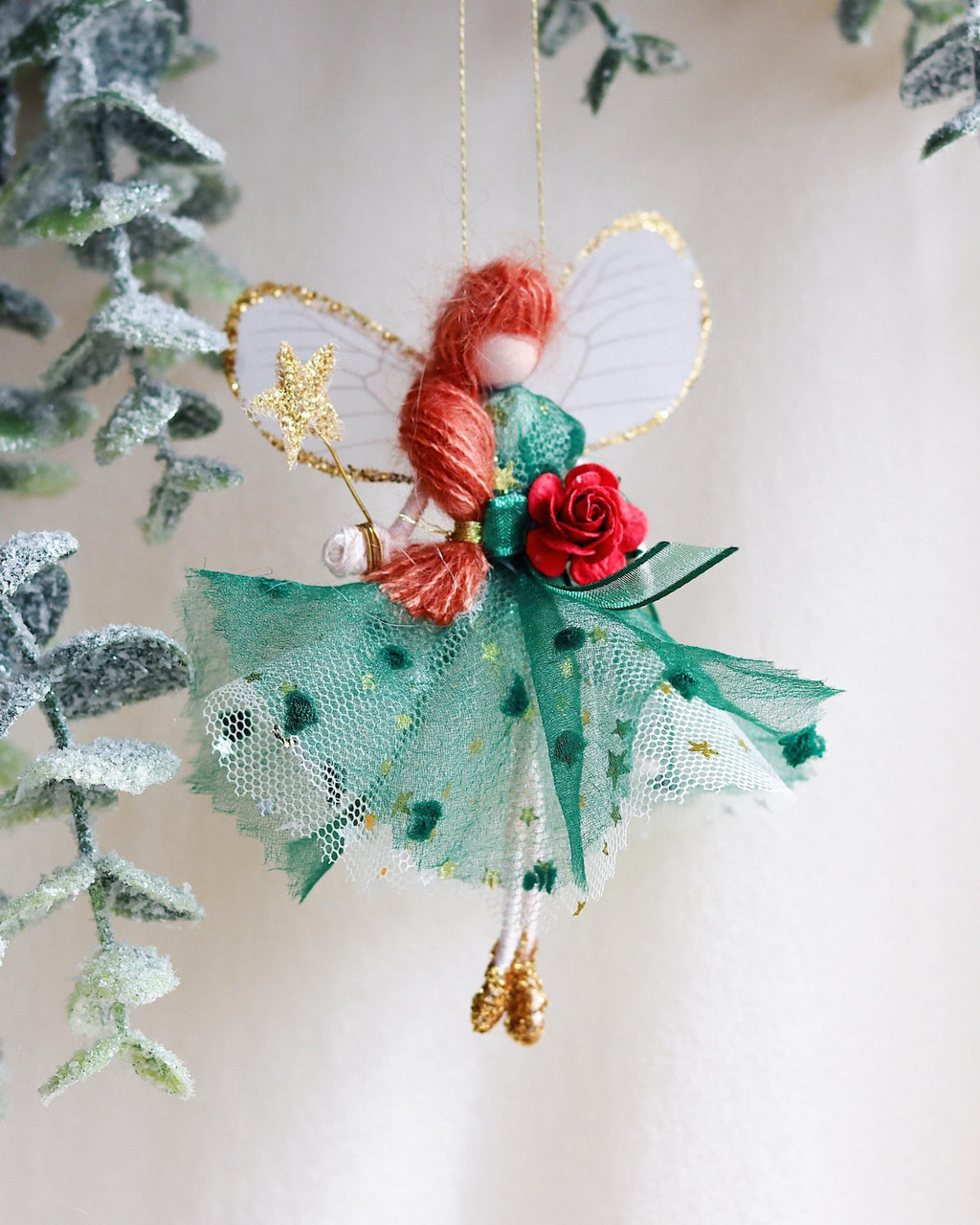 Christmas Decorative fairy ornament with red hair and a red rose, hanging against a blurred green leafy background.