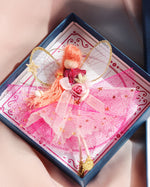 Load image into Gallery viewer, A small handmade fairy ornament with pink wings, ginger hair, and a pink dress sits inside an open navy gift box lined with pink patterned paper. A branded card and part of a navy box with a gold circular logo are visible beside it.
