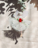 Load image into Gallery viewer, A handmade fabric angel ornament hanging from a string, featuring white wings, a white tulle dress with small blue and black dots, a red fabric rose at the waist, and a pale green ribbon, set against a beige background with leaf shadows.
