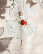 Load image into Gallery viewer, A delicate fairy ornament with translucent white wings, a white tulle dress with small blue and green sequins, a red fabric flower at the waist, and a green ribbon, hanging against a soft beige background with leaf shadows.
