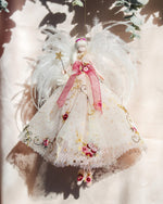 Load image into Gallery viewer, A decorative angel ornament hanging against a light fabric backdrop with leaf shadows. The ornament has white feather wings, a white embroidered dress with pink ribbon accents, floral details, and a small gold star wand.
