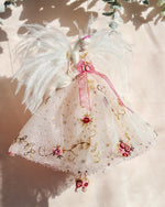 Load image into Gallery viewer, A decorative angel ornament with white feathers, a white tulle dress with gold embroidery, and pink ribbon accents, hanging against a warm beige background with soft shadows and greenery.
