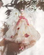 Load image into Gallery viewer, A decorative angel ornament hanging from a string, featuring white feather wings, a pink ribbon bow, and a sheer cream dress with gold glitter and embroidered floral details, set against a bright white background with leaf shadows.
