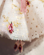 Load image into Gallery viewer, Close-up of a white tulle tutu skirt with gold embroidered stars and swirls, pink rose motifs, a pink ribbon, and small pink-and-white ballet shoes hanging below.
