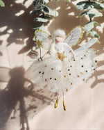 Load image into Gallery viewer, A white fairy Christmas ornament with gold glitter stars, translucent wings, and a sparkly white tulle skirt, hanging against a beige background with soft shadows from green leaves.
