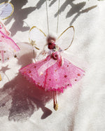 Load image into Gallery viewer, A pink fairy ornament with gold star details, translucent wings, and a small rose on its head, hanging against a white textured background with soft shadows. A second similar ornament is partially visible at the upper left.
