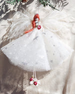 Load image into Gallery viewer, A handmade angel Christmas ornament with a white tulle dress decorated with silver glitter stars, a red felt hair bun, and red berries, hanging from a pine branch against a textured beige fabric background.
