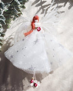 Load image into Gallery viewer, A white angel Christmas ornament with silver glitter star details, red hair, and a small red berry accent, hanging against a textured beige fabric background with soft shadows and greenery.
