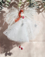 Load image into Gallery viewer, A white angel Christmas ornament with silver glittery star details, a red-haired figure, and small red berries hanging from the skirt, shown against a warm beige background with soft shadows.
