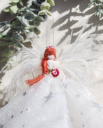 Load image into Gallery viewer, A decorative angel ornament with white feather wings, a white tulle dress with silver star details, and bright orange hair, hanging against a softly lit snowy background with frosted greenery.

