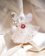 Load image into Gallery viewer, A delicate fairy ornament with white wings trimmed in gold glitter, a white tulle dress with gold star details, and a pink rose at the center, hanging against a draped cream satin background.
